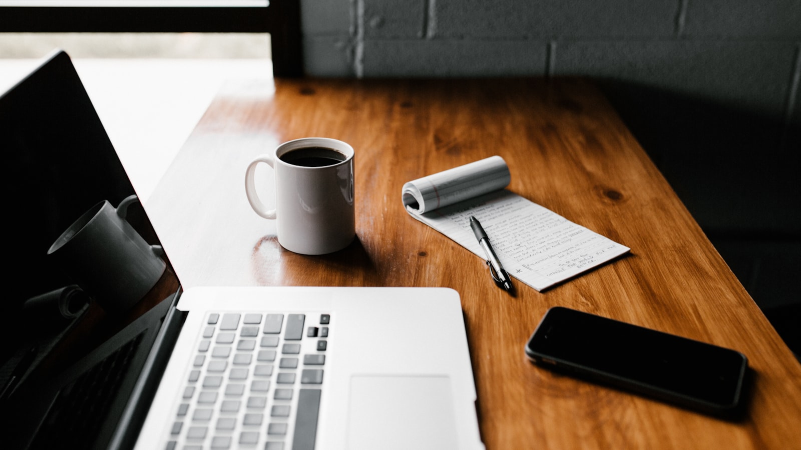 An open notebook and a cup of coffee on a wooden desk by a window in soft morning light.